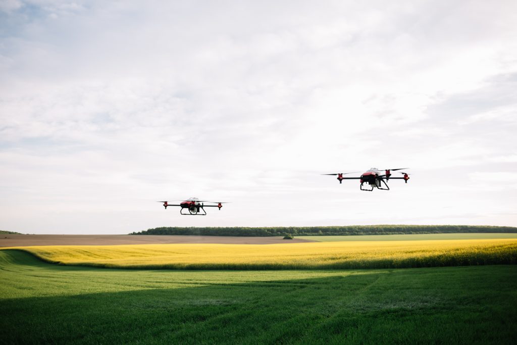 Two drones soar above a green and yellow crop field under a bright, cloudy sky, showcasing the skills gained through RPAS Training. Distant trees and rolling hills complete the picturesque backdrop.