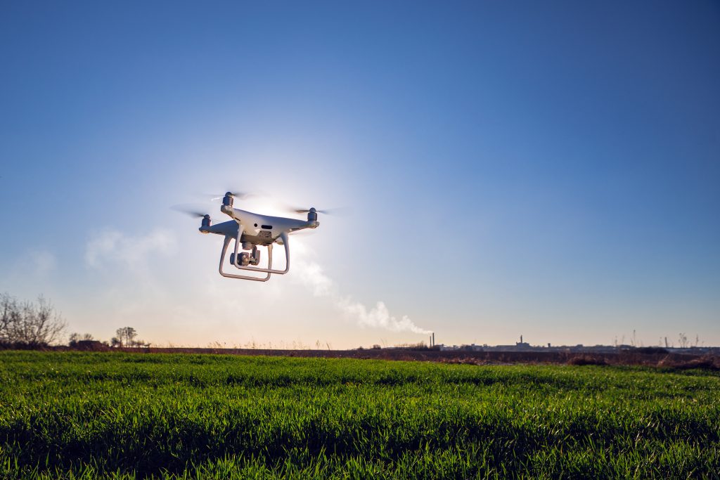 A white drone hovers above a green grassy field under a clear blue sky, sunlight behind it and a distant city skyline on the horizon—a perfect scene for hands-on RPAS Training with Drone Training Australia.