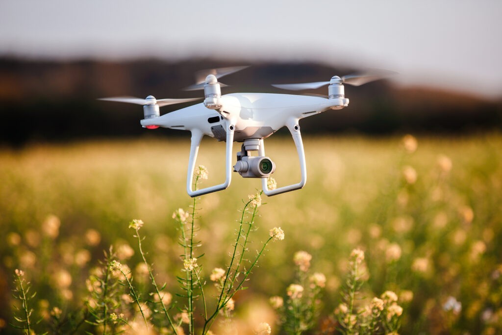 A white quadcopter drone with a camera, perfect for UAV training, hovers above a field of yellow wildflowers, with a blurred natural landscape in the background.