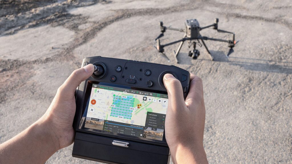 A person participates in in-person drone training, holding a remote controller with a screen displaying a map and controls, operating a professional drone on a rough outdoor surface.