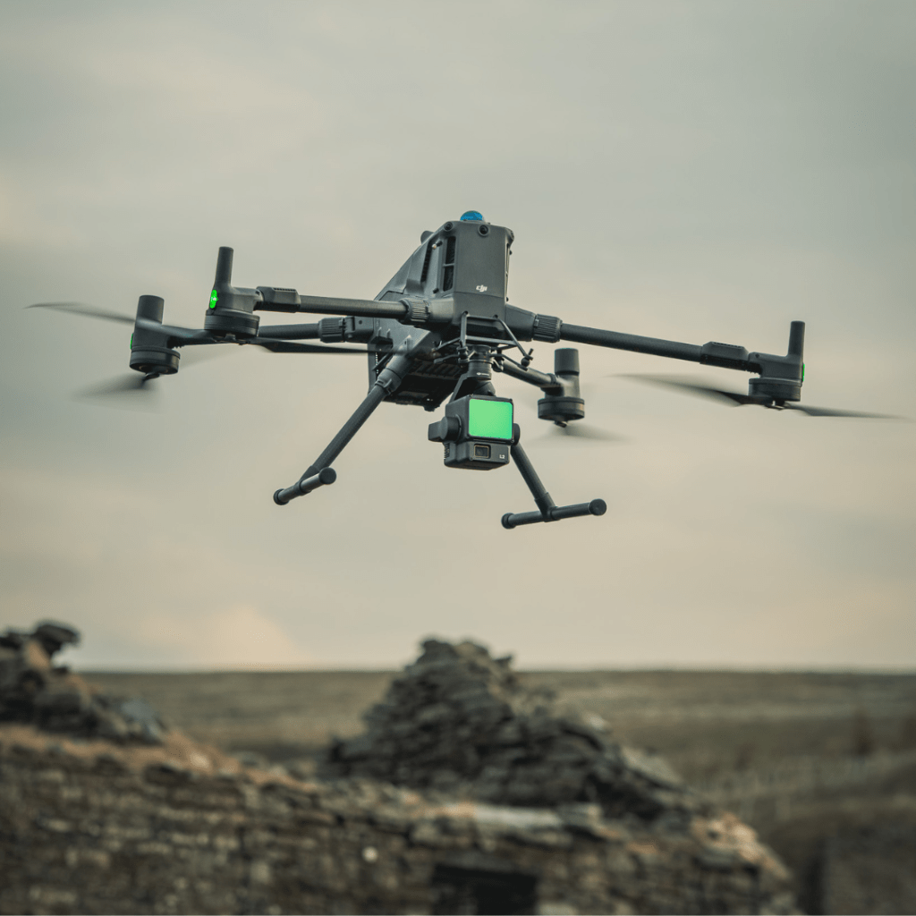 A black quadcopter drone with a mounted camera hovers in the air above rocky ruins, showcasing how drones can assist in land management amid a blurred landscape and cloudy sky.