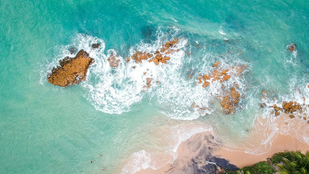 Aerial view captured by drones shows turquoise ocean waves crashing over rocky formations and a sandy shoreline, with some greenery visible at the bottom edge—ideal for training in landscape photography.